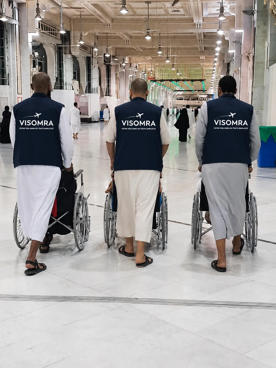 Trois accompagnateurs Visomra, en gilet floqué, poussant des pèlerins en fauteuil roulant dans les galeries de la Grande Mosquée de La Mecque.